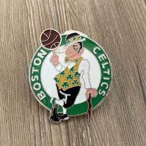 May include: A green, white, and silver Boston Celtics pin featuring a cartoon leprechaun holding a basketball and a cane. The pin says "Boston Celtics" around the outside.