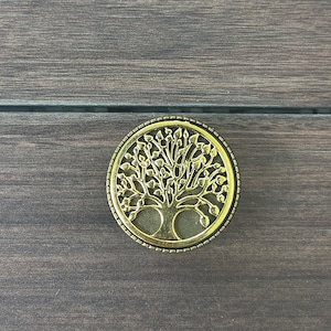 May include: A gold-colored metal ornament with a tree of life design. The ornament is round and has a detailed design with leaves and branches.