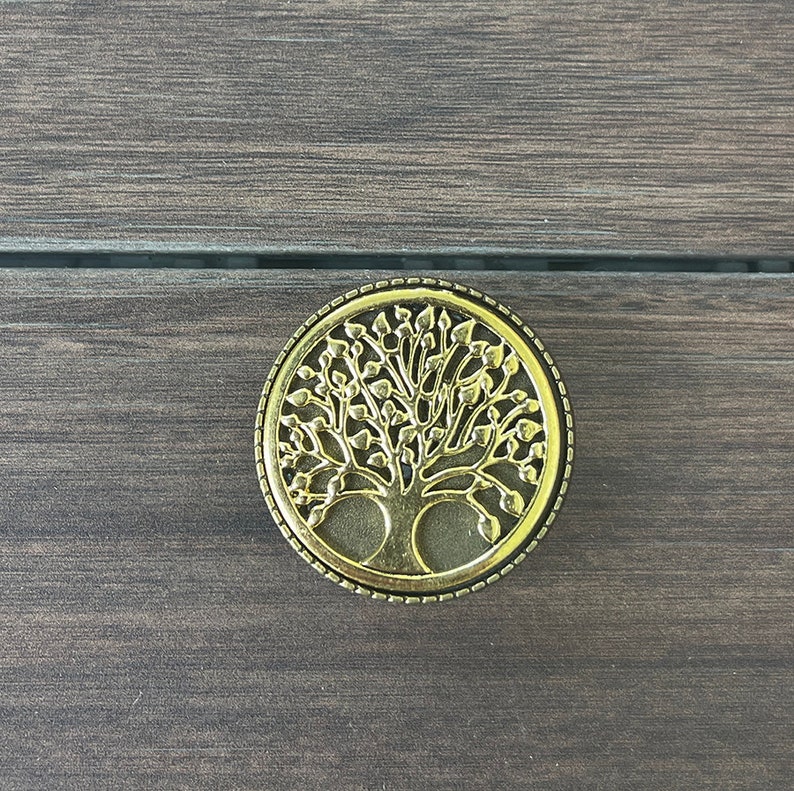 May include: A gold-colored metal ornament with a tree of life design. The ornament is round and has a detailed design with leaves and branches.