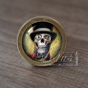 Pode incluir: Um botão de armário redondo de cor latão com um inserto de vidro que apresenta uma caveira usando um chapéu-coco e um monóculo. A caveira está segurando um cachimbo e tem uma gravata borboleta.