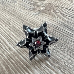 Puede incluir: Un colgante de estrella de seis puntas de plata con una piedra preciosa roja en el centro. La estrella tiene detalles intrincados y un acabado negro.