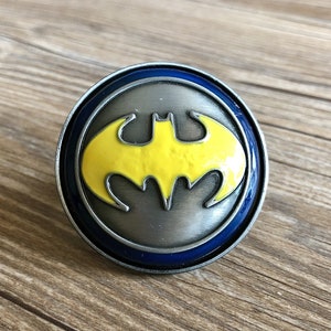 Może przedstawiać: Srebrna klamra do paska z niebieskim obramowaniem i żółtym logo Batmana.