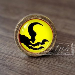 May include: A round cabinet knob with an antique brass finish and a domed glass insert. The insert features a bright yellow background with a black silhouette graphic of a sharp, curved claw or talon reaching across the center of the circle.