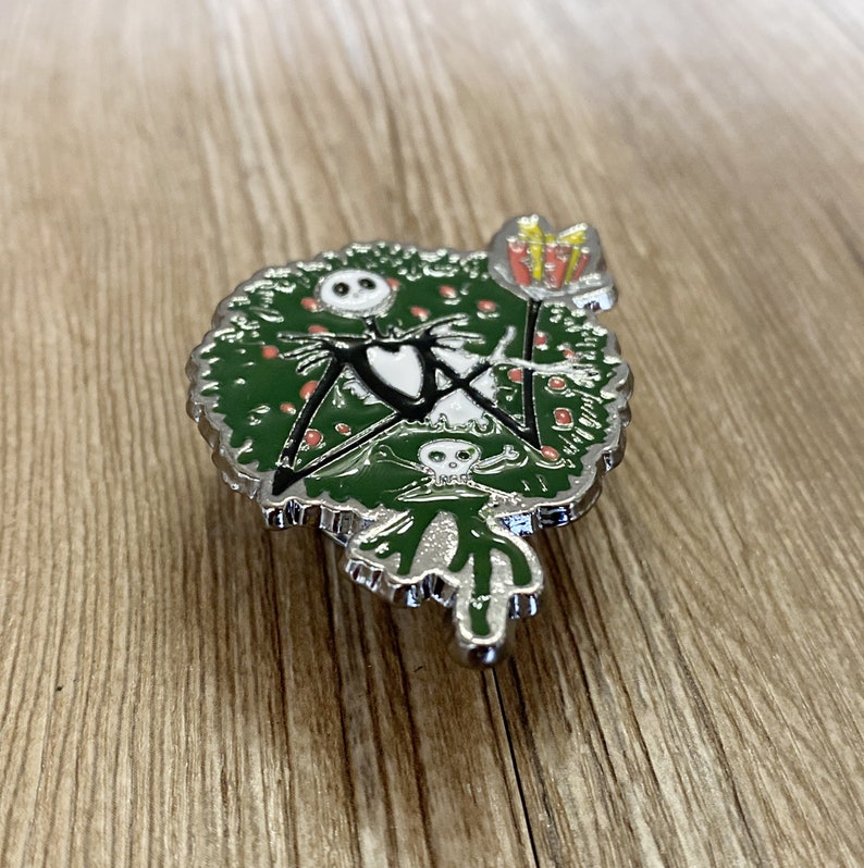 以下が含まれることがあります： 緑色のクリスマスツリーのピンで、白と黒のジャック・スケリントンが座っています。ツリーは赤いオーナメントと黄色の星で飾られています。