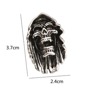 Op de afbeelding: Een zilverkleurige schedelring met een ontwerp van een Grim Reaper. De ring is 3,7 cm hoog en 2,4 cm breed. De schedel heeft een open mond en draagt een kap. De ring is waarschijnlijk gemaakt van metaal.