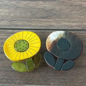 May include: Two sunflower-shaped brooches. One is yellow with a green center and green leaves. The other is brown with a dark center and black leaves. Both are on a wooden surface.