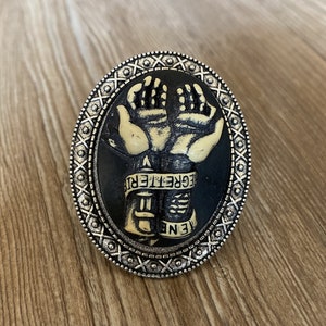 May include: A black cameo ring with a silver-toned setting. The cameo features two hands clasped together with the text "Egregelter" on a band.