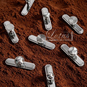 May include: A collection of shiny, silver cabinet pulls with various knob designs. The pulls are rectangular with decorative edges and are arranged on a textured, brown surface. The image includes the text "Lotus Hardware".