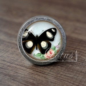 May include: A silver knob with a glass insert featuring a black butterfly with white markings against a white background with pink and green floral accents.