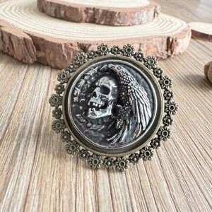 May include: A round, antique-style brooch with a skull and raven design. The central image is surrounded by a floral border. The brooch has a silver and bronze color scheme, with intricate detailing.