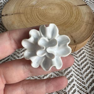 Könnte beinhalten: Eine weiße Keramikblume mit geschichteten, abgerundeten Blütenblättern und einer glänzenden Oberfläche. Das Objekt zeigt eine zentrale Anordnung kleinerer, geschwungener Blütenblätter, die von einem äußeren Ring aus größeren Blütenblättern umgeben ist.