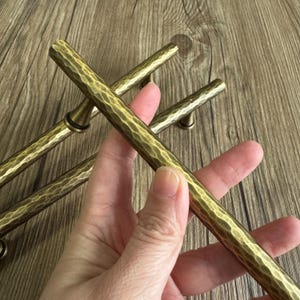 Peut inclure: Gros plan sur plusieurs poignées de meubles en laiton antique avec une texture martelée. Les poignées sont cylindriques avec un aspect légèrement vieilli, suggérant une esthétique vintage ou rustique. La couleur laiton chaud contraste avec la surface en bois.
