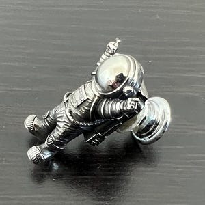 May include: A silver astronaut figurine with a raised arm and a round, reflective helmet. The astronaut is holding a silver disc in its other hand.