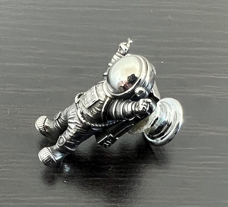 May include: A silver astronaut figurine with a raised arm and a round, reflective helmet. The astronaut is holding a silver disc in its other hand.