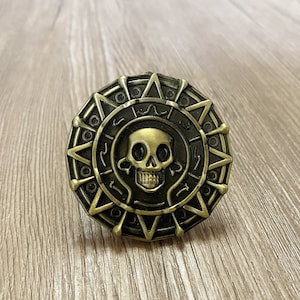 May include: A gold-colored metal pirate coin with a skull and crossbones design. The coin has a raised edge and a textured surface.