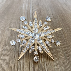 May include: A gold-toned star-shaped brooch with clear rhinestones. The brooch has a pointed star shape with multiple points and is decorated with clear rhinestones of varying sizes.