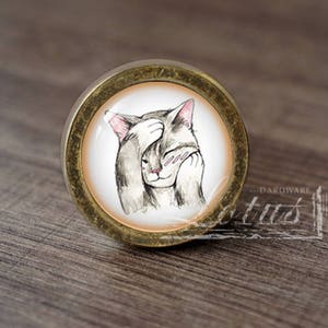 May include: A round, gold-coloured cabinet knob with a white background and a cartoon image of a cat covering its eyes with its paw. The cat has grey fur and pink ears.