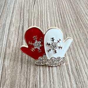 Könnte beinhalten: Ein Paar rote und weiße Fäustlinge mit Strass-Schneeflocken. Die Fäustlinge sind miteinander verbunden und haben eine goldene Oberfläche.