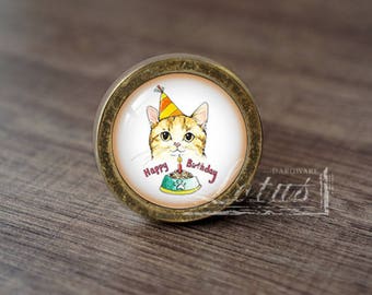 May include: A gold-toned round cabinet knob with a glass insert featuring a cartoon cat wearing a party hat and holding a birthday cake. The text "Happy Birthday" is printed on the glass insert.