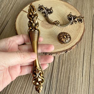 以下が含まれることがあります： 装飾的なアンティーク真鍮金具セット。長いハンドル、2つの小さなハンドル、丸いノブが含まれています。 家具の修復や装飾プロジェクトに最適な、詳細な花柄とスクロールワークのデザインが特徴です。