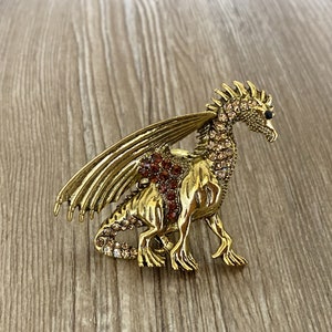May include: A gold-toned dragon brooch with brown and clear rhinestones. The dragon has wings and a long tail.