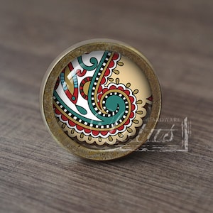 May include: A round, antique brass cabinet knob with a colourful paisley design. The knob has a glass top with a beige background and a teal, red, and gold paisley pattern.