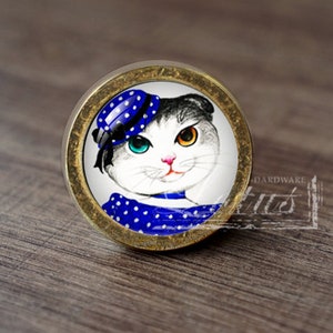 May include: A round, gold-toned cabinet knob with a white porcelain insert featuring a cartoon cat wearing a blue polka dot dress and hat. The cat has one blue eye and one brown eye.