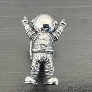 May include: A silver astronaut figurine with raised arms and a shiny helmet. The figurine is standing on a grey surface.