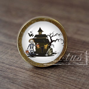 Pode incluir: Um botão de armário redondo de cor dourada com um fundo branco e um design temático de Halloween. Um esqueleto de desenho animado está segurando um balde de abóbora e caminhando em direção a uma casa mal-assombrada com um morcego voando acima.