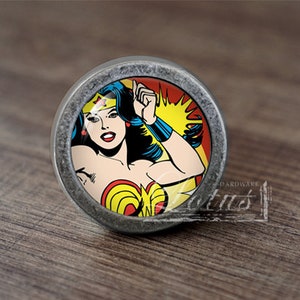 May include: A round, silver-colored cabinet knob with a colorful image of Wonder Woman in a red, yellow, and blue costume. The background is red and yellow.