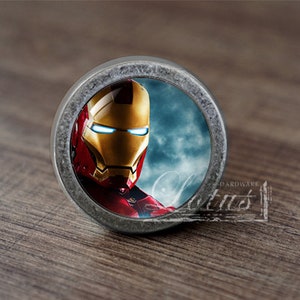 May include: A round, metallic Iron Man-themed object. The image features the superhero's helmet in gold and red, with blue eyes, set against a cloudy sky background. The object has a distressed metal border and a small, rectangular tag with the word "Hardware".