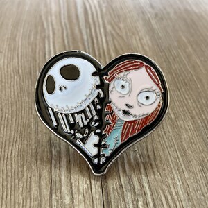 May include: A black heart-shaped enamel pin with a white and black Jack Skellington character on the left side and a red-haired Sally character on the right side.
