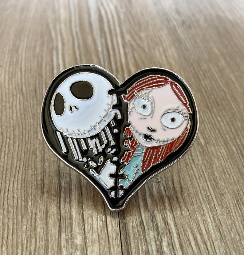 May include: A black heart-shaped enamel pin with a white and black Jack Skellington character on the left side and a red-haired Sally character on the right side.