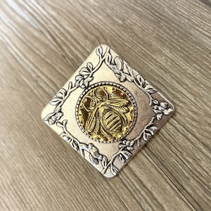 May include: A silver square pendant with a gold bee design in the center. The bee is surrounded by a floral design.