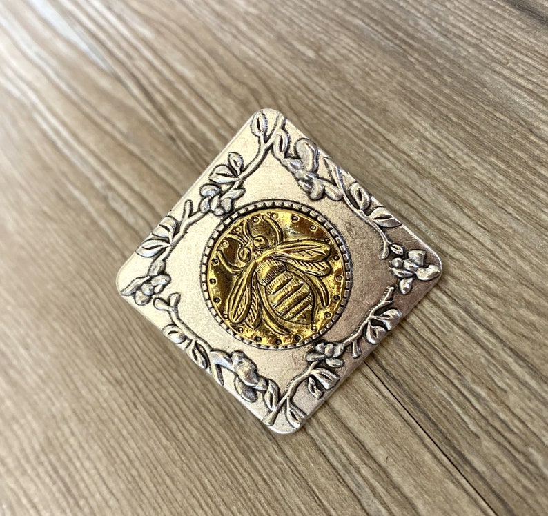 May include: A silver square pendant with a gold bee design in the center. The bee is surrounded by a floral design.