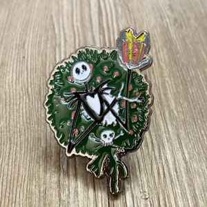 以下が含まれることがあります： ナイトメア・ビフォア・クリスマスのジャック・スケリントンを描いた緑と赤のエナメルピン。ジャックはプレゼントを持って、リースの前に立っています。