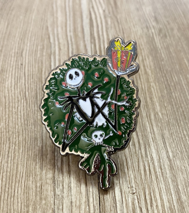 以下が含まれることがあります： ナイトメア・ビフォア・クリスマスのジャック・スケリントンを描いた緑と赤のエナメルピン。ジャックはプレゼントを持って、リースの前に立っています。