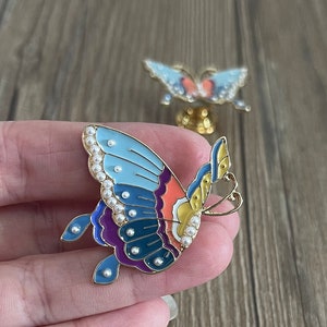 May include: A colourful butterfly brooch with a blue, purple, orange and yellow enamel design. The butterfly has a gold finish and is decorated with white pearl accents.