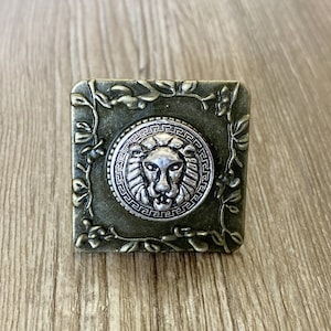 May include: A square metal button featuring a lion's head design in the centre. The button is surrounded by a decorative floral border and has a silver finish.