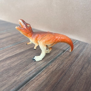 May include: A small, orange and cream-coloured Tyrannosaurus Rex toy dinosaur with an open mouth, revealing white teeth. The toy has a curved tail and is posed on a wooden surface.
