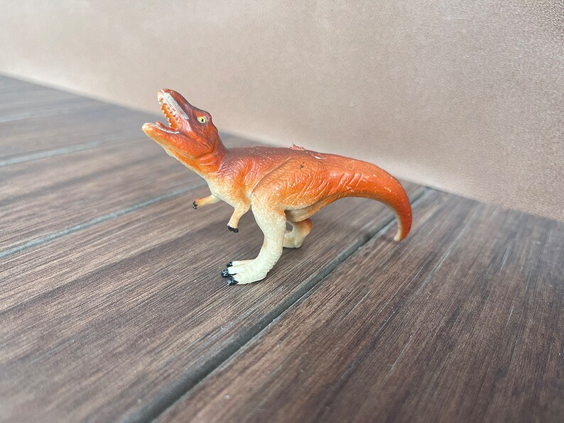 May include: A small, orange and cream-coloured Tyrannosaurus Rex toy dinosaur with an open mouth, revealing white teeth. The toy has a curved tail and is posed on a wooden surface.