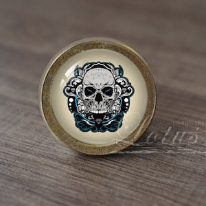 Op de afbeelding: Een ronde, antieke messing kastknop met een glazen inzetstuk met een witte schedel met blauwe accenten en ingewikkelde zwarte bloemmotieven.