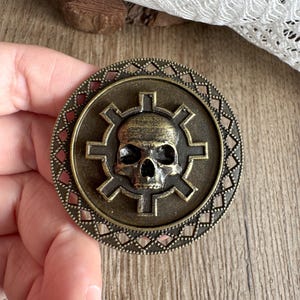 Op de afbeelding: Een ronde, antiek messingkleurige metalen hanger met een schedel in het midden, omringd door een tandwielachtig ontwerp. De buitenrand heeft een decoratief patroon met kleine, ruitvormige inzetstukken. De hanger wordt in een hand gehouden.