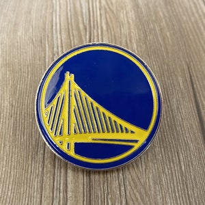 May include: A blue and yellow enamel pin featuring the Golden State Warriors logo, a stylised depiction of the Golden Gate Bridge.