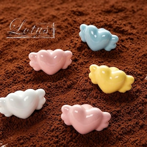 Puede incluir: Cinco piezas decorativas de cerámica en forma de corazón en blanco, rosa, azul claro y amarillo, cada una con detalles en forma de alas. Las piezas están dispuestas sobre una superficie marrón texturizada. La palabra "Lotus" es visible en la esquina superior izquierda.
