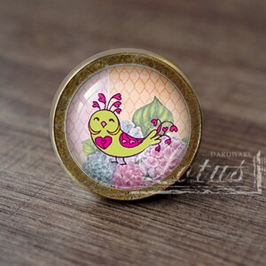 May include: A round, brass-colored cabinet knob with a glass insert featuring a green bird with pink hearts on a floral background.