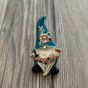 May include: A blue and gold gnome figurine with a white beard and a floral hat. The gnome is holding a wreath of flowers in its hands.