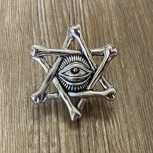 Peut inclure: Pendentif argenté en forme d'étoile à six branches avec un œil qui voit tout. L'étoile est composée de pièces en forme d'os.