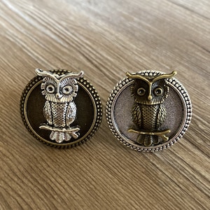 May include: Two round, silver and bronze owl-shaped buttons with a detailed design. The owls are perched on a branch with their wings spread.