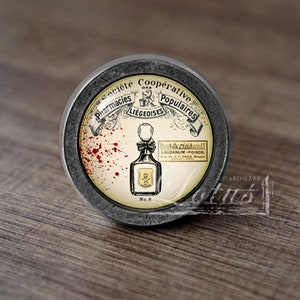 Könnte beinhalten: Ein runder, silberfarbener Schrankknopf mit einem Glaseinsatz. Der Glaseinsatz zeigt eine Vintage-Illustration einer Flasche mit Totenkopf und Knochen und dem Text "Laudanum - Poison". Der umgebende Text lautet "Societe Cooperative Pharmacies Liegeoises Populaires".